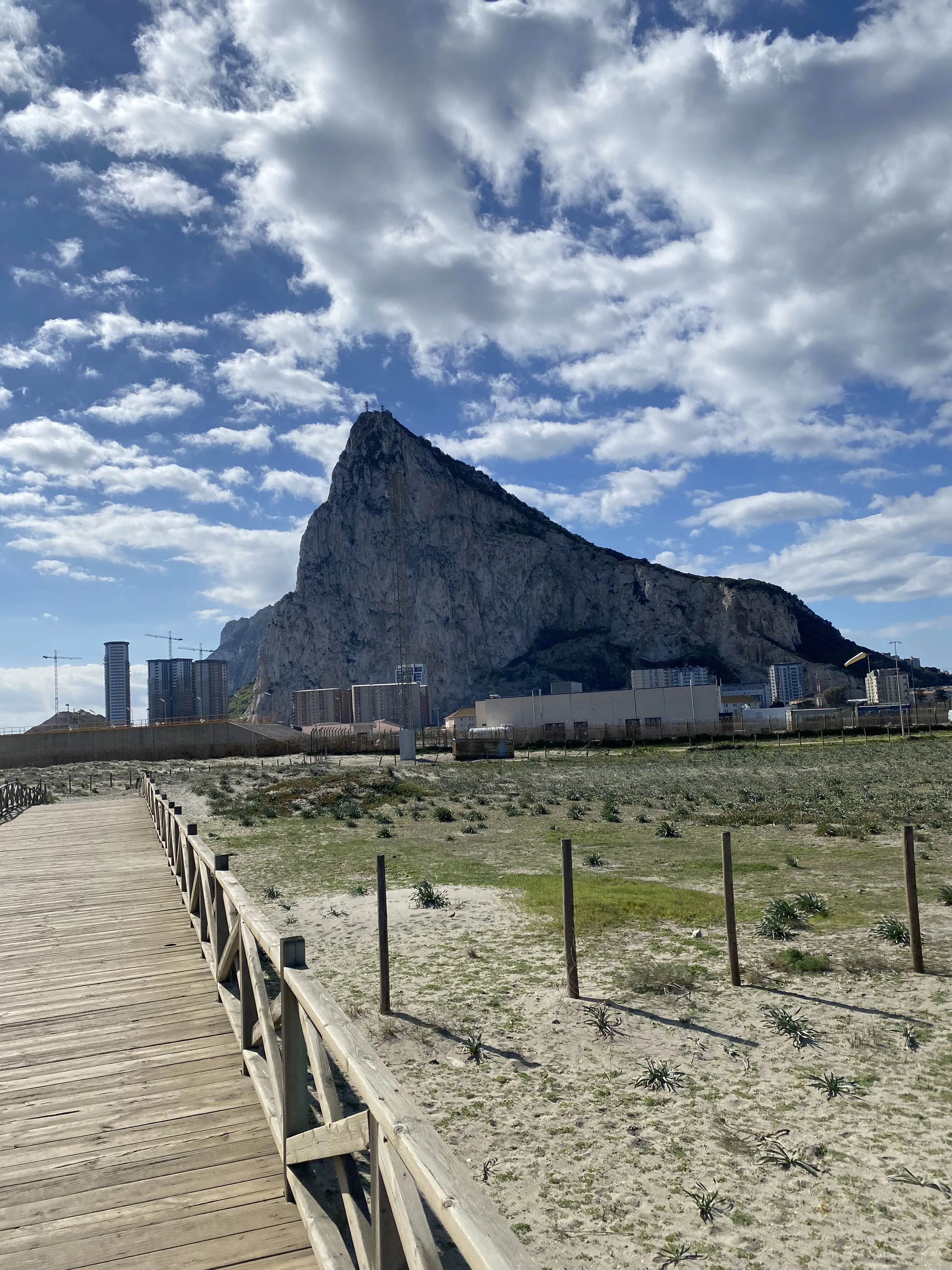 A boardwalk over dunes running towards the Rock of Gibraltar under broken cloud.