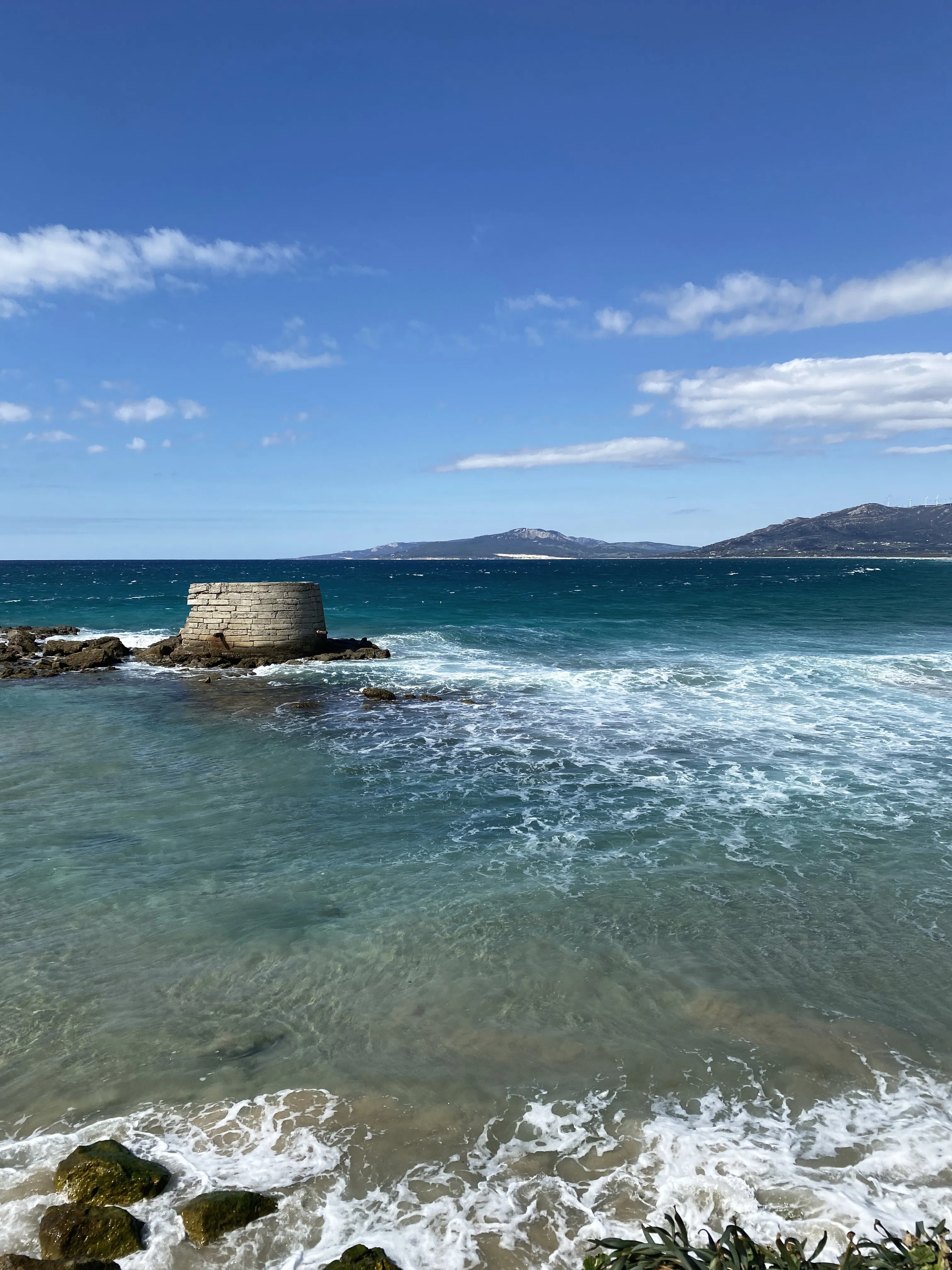 A ruined stone jetty standing in breaking surf, a headland across the water.