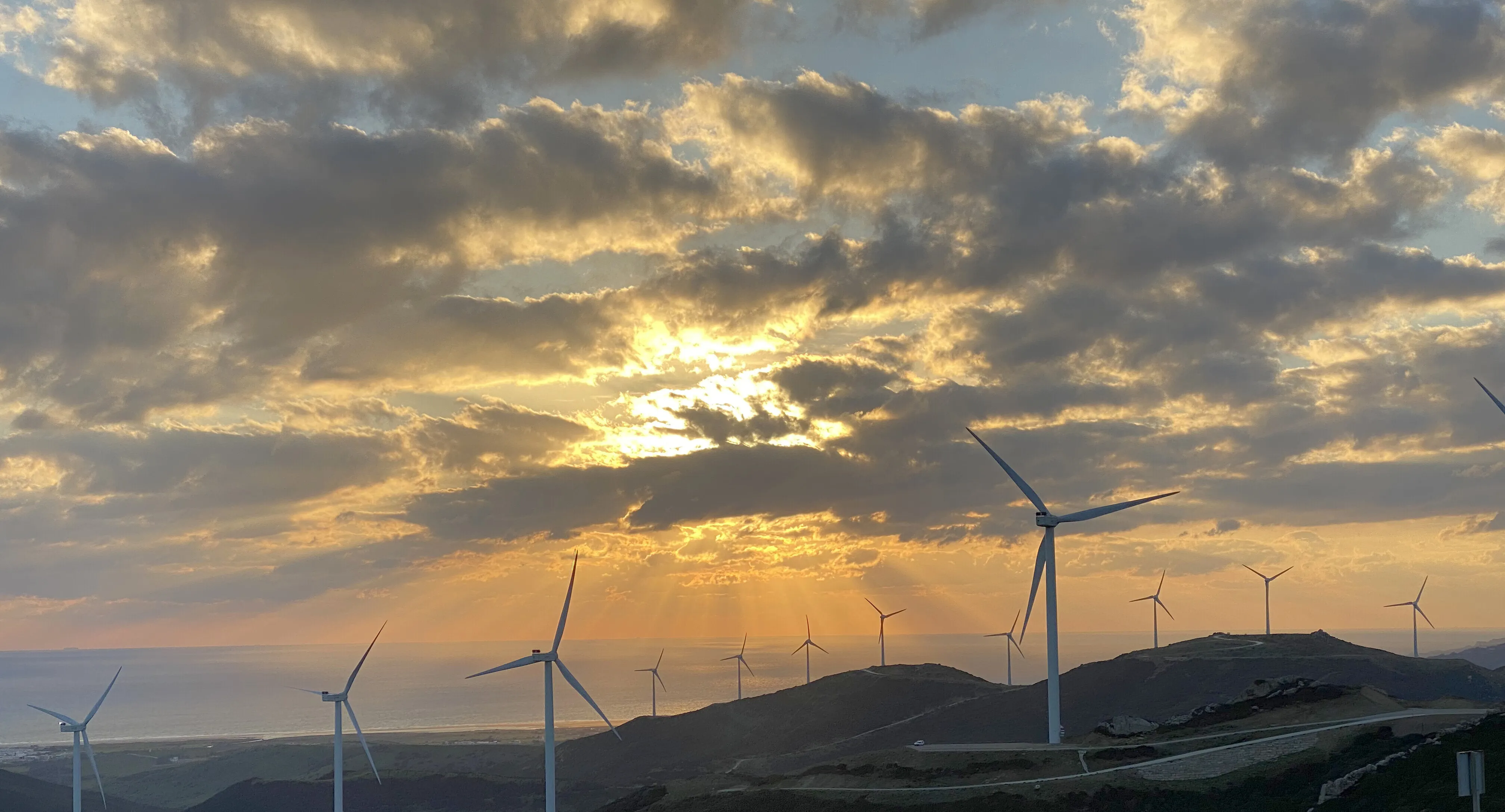 Wind turbines along a ridge with the sun breaking through cloud behind them.